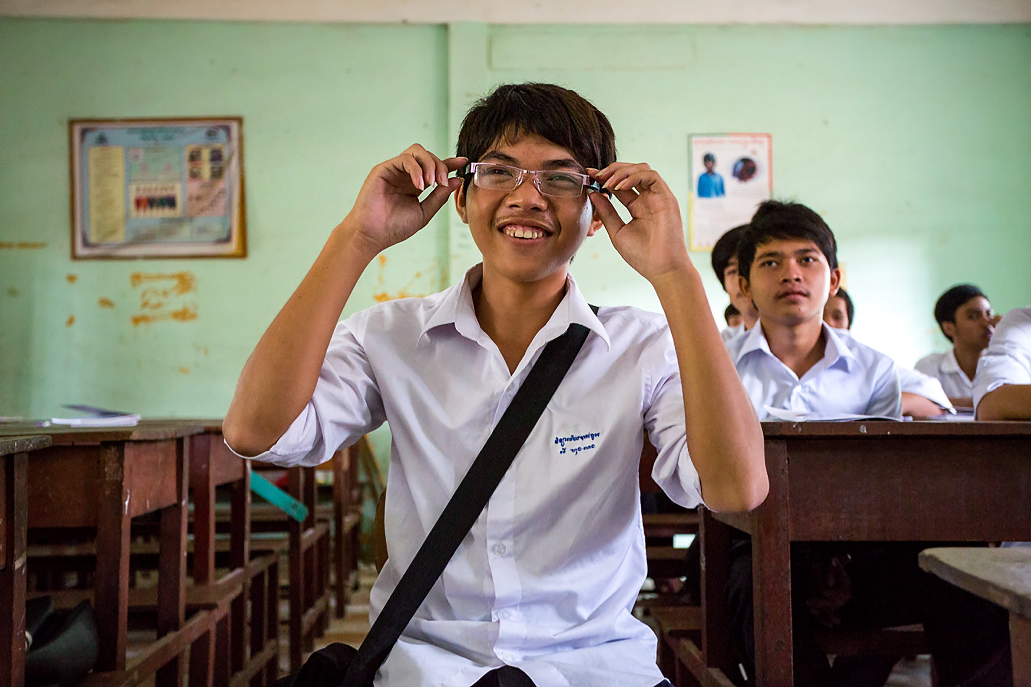 A boy in a classroom smiles as he puts on glasses