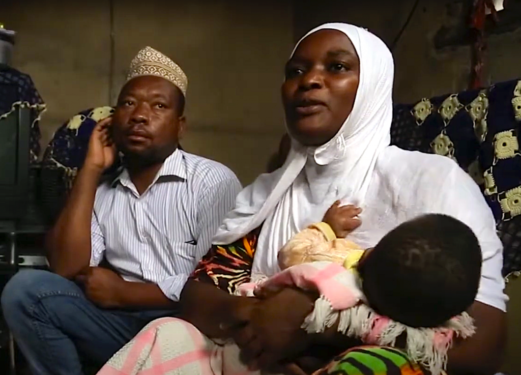 A man and woman sit next to each other. The woman holds a baby in her arms. She wears a white headdress 