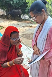 In India, a woman holds a phone and a clipboard in front of a seated woman. There is a piece of string between them