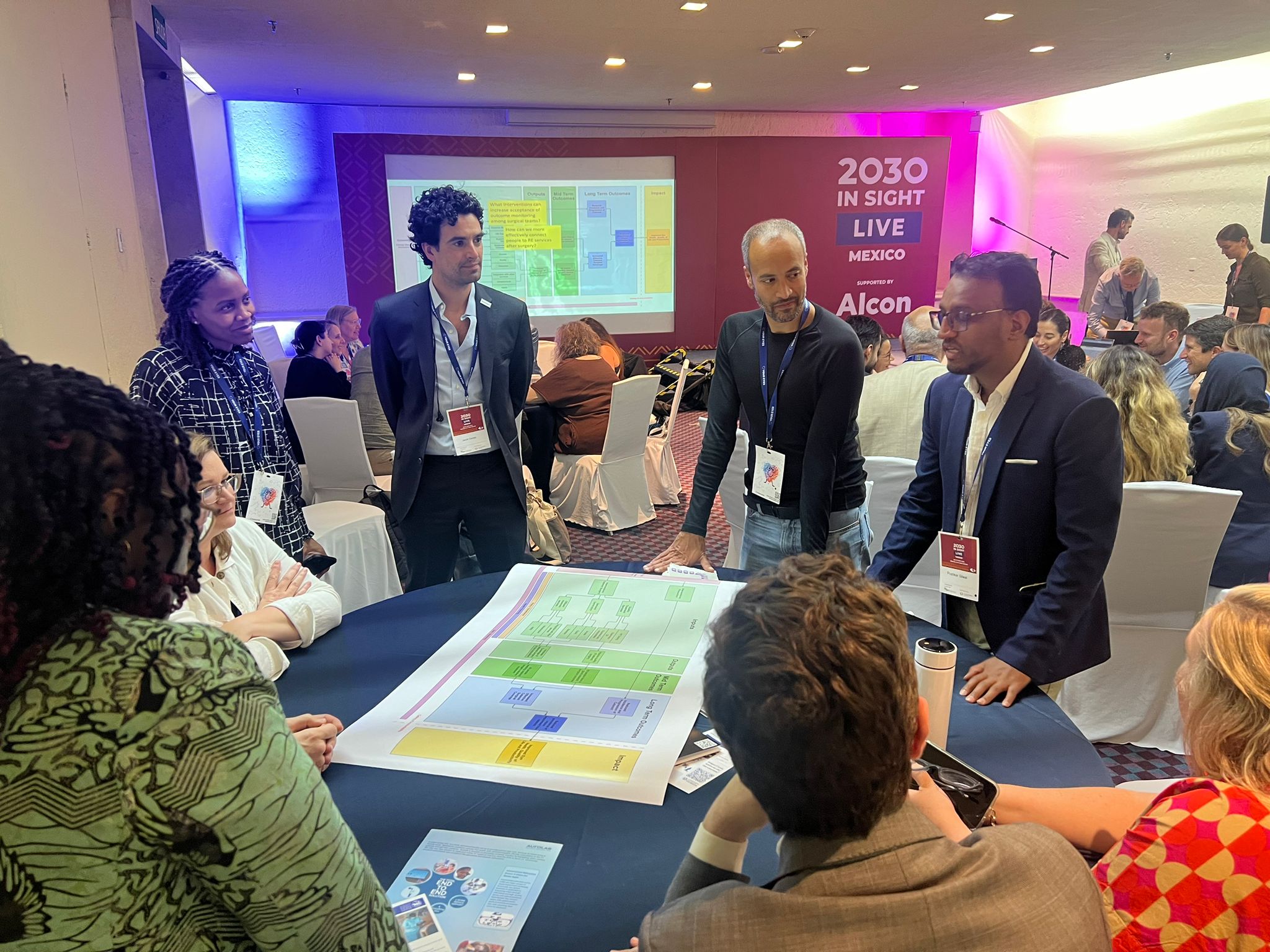a group of people stand around a table at a conference centre, looking at a large process diagram