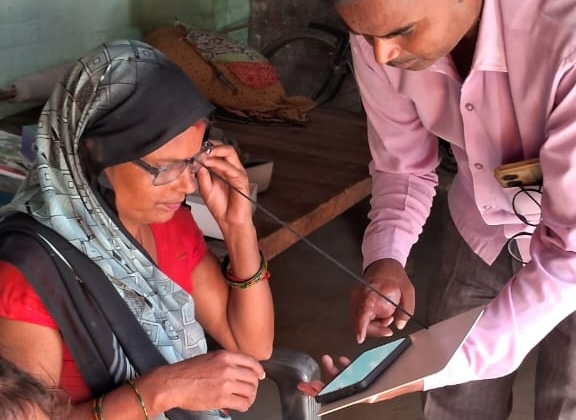 In india, a health worker holds a phone to test a woman's vision who is seated wearing glasses