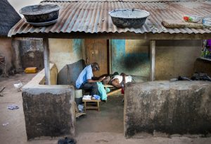 outside a house, a man lies down as another performs trachoma surgery on him