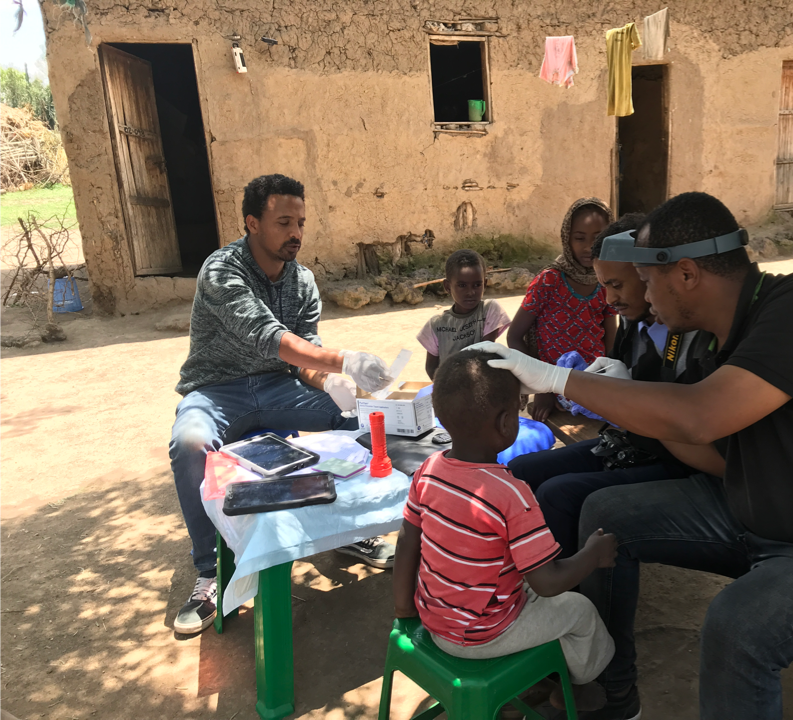 In Ethiopia, two healthcare researchers swab children's eyelids at a table outside