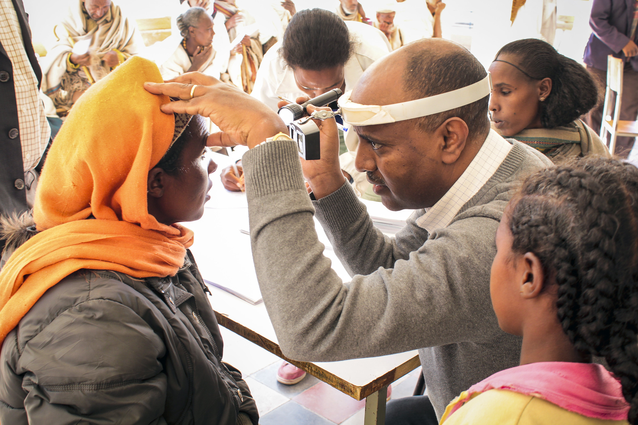 a health worker holds up a torch to a woman in an orange headress' eye