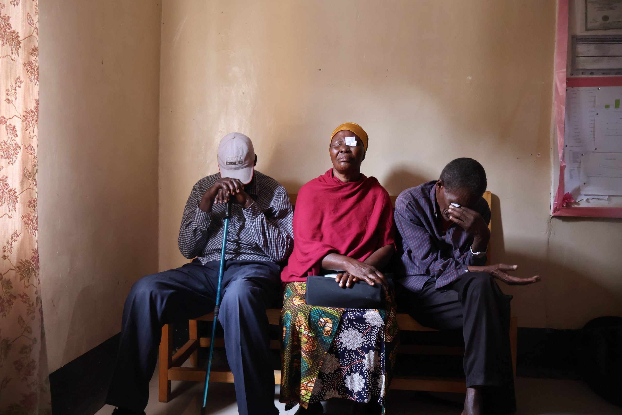in a darkened room in Tanzania, three people sit in a row covering their eyes. One woman has an eye patch